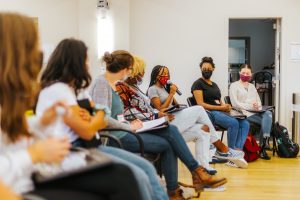 A group of facilitators in facilitator training is seated in a large circle wearing masks. One facilitator holds a microphone and the others pictured are looking at her.